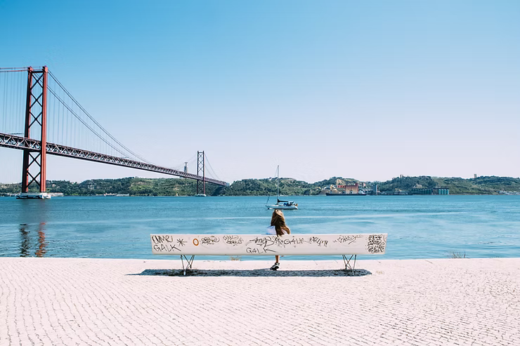 View of the Tejo river in Lisbon with the 25th April bridge on the left and a person sitting on a bench in the centre
